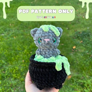 Peut inclure: Un chat au crochet fait à la main dans un chaudron noir avec de la slime verte. Le chat gris a des yeux verts et un nez rose. Le chaudron est noir et repose sur un fond herbeux vert. Le texte "PDF PATTERN ONLY" est en haut.