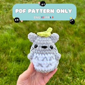 May include: A crocheted gray and white Totoro amigurumi plush toy with a green leaf on its head. The toy is being held in a hand against a green grass background. The text "PDF PATTERN ONLY ENG/FR" is displayed above the toy.