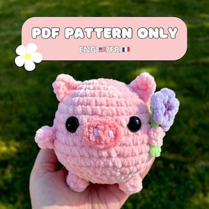 Peut inclure: Un cochon rose en crochet tenant une fleur violette. Le cochon a des yeux noirs et un groin rose. Le cochon est assis sur une main.