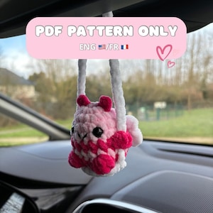 Könnte beinhalten: Ein rosa und weiß gehäkeltes Amigurumi, das an einem Rückspiegel hängt. Das Amigurumi hat die Form eines Eichhörnchens und hält sich an einer weißen Seilschaukel fest. Der Text "PDF PATTERN ONLY ENG/FR" ist auf dem Bild zu sehen.