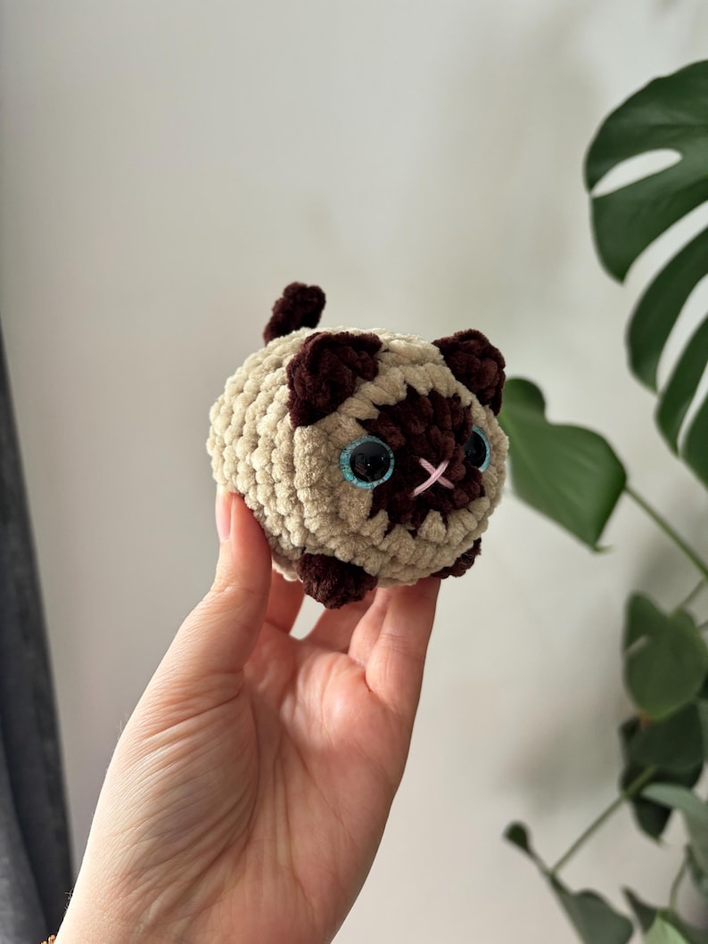 May include: A crocheted cat toy in a Siamese cat design. The plush toy features a beige body with dark brown accents on the ears, face, and paws. It has blue eyes and a pink embroidered nose. The cat toy is round and soft.
