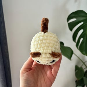 May include: A handmade, cream-colored crochet doll with brown accents. The doll has small, dark eyes and a brown, cylindrical top. The doll is round and appears to be a small, plush toy. The background is a plain wall and a green plant.