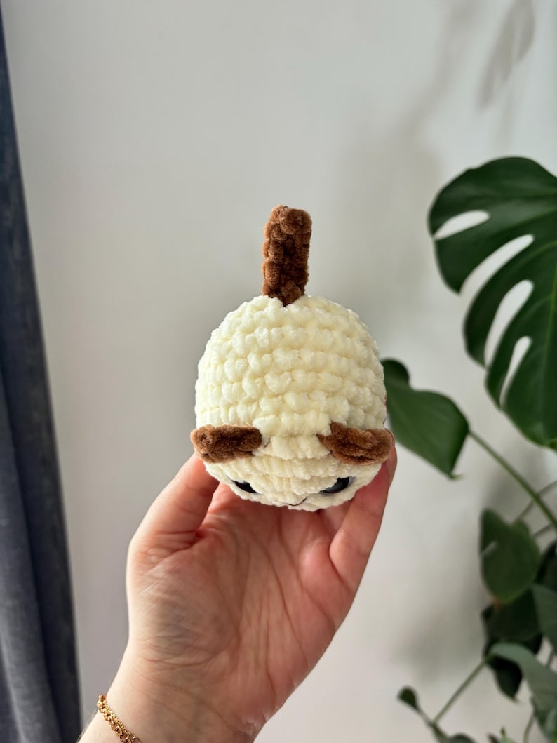 May include: A handmade, cream-colored crochet doll with brown accents. The doll has small, dark eyes and a brown, cylindrical top. The doll is round and appears to be a small, plush toy. The background is a plain wall and a green plant.
