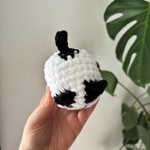 May include: A handmade, crocheted panda bear toy. The panda is white with black accents on the ears and eyes, and a black stem. The toy is held in a hand, with a gold bracelet visible. The background is a white wall and green plant.