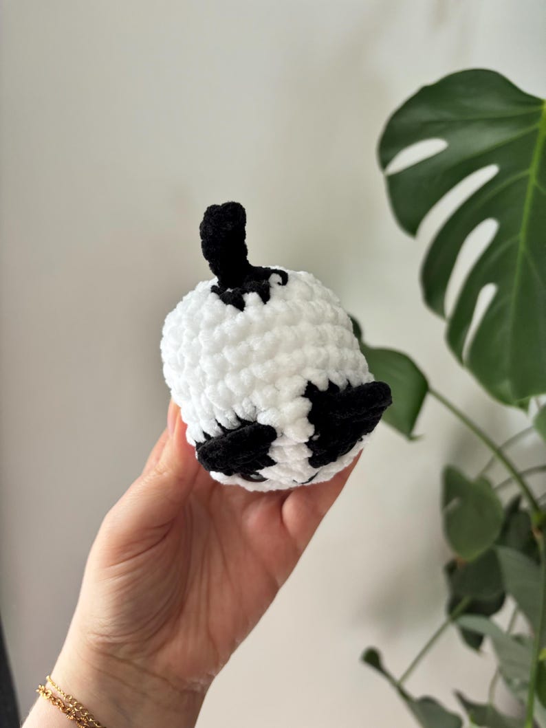 May include: A handmade, crocheted panda bear toy. The panda is white with black accents on the ears and eyes, and a black stem. The toy is held in a hand, with a gold bracelet visible. The background is a white wall and green plant.