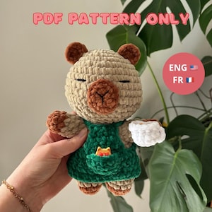 Peut inclure: Une poupée ours au crochet portant un tablier vert avec une poche et tenant une tasse blanche. L'ours est beige avec des accents marron et les yeux fermés. Le texte "PDF PATTERN ONLY" est en haut de l'image.
