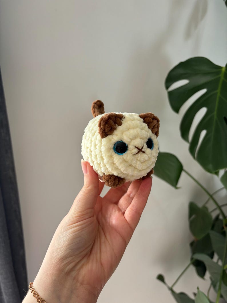 May include: A small, round, cream-colored crocheted cat with brown accents on the ears, paws, and tail. The cat has large blue eyes and a stitched nose. The handmade plush toy is held in a hand.