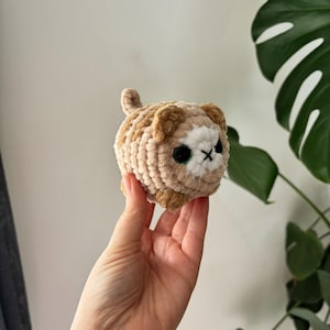 May include: A crocheted cat toy in shades of tan, brown, and white. The handmade plushie has black eyes and a stitched 'X' for a mouth. The cat has a small tail and is held in a hand.
