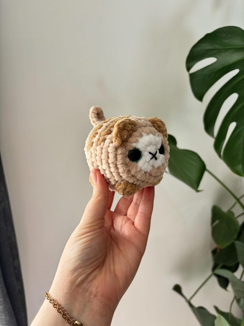 May include: A crocheted cat toy in shades of tan, brown, and white. The handmade plushie has black eyes and a stitched 'X' for a mouth. The cat has a small tail and is held in a hand.