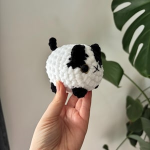 May include: A handmade, plush, round cat toy in white with black patches. The cat has black ears, paws, and a tail. The eyes are small, black beads, and the mouth is an embroidered X. The toy is held in a hand.