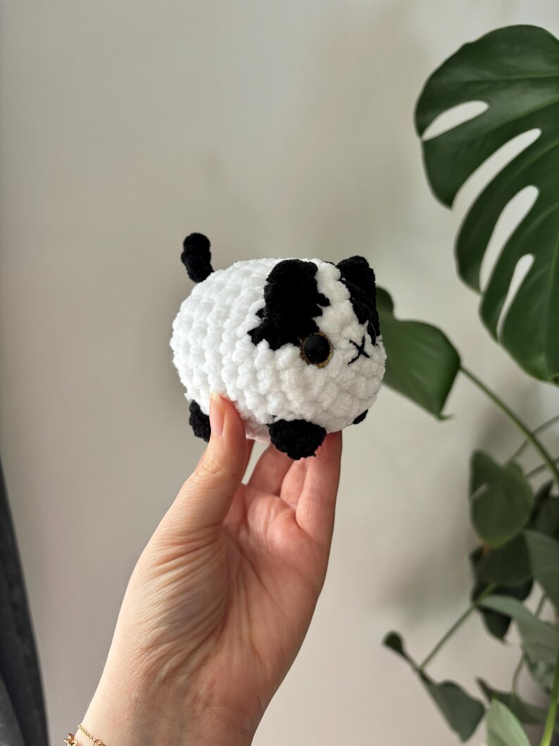 May include: A handmade, plush, round cat toy in white with black patches. The cat has black ears, paws, and a tail. The eyes are small, black beads, and the mouth is an embroidered X. The toy is held in a hand.