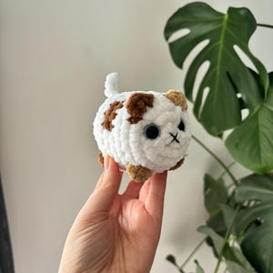May include: A handmade, white crocheted cat with brown spots, blue eyes, and a stitched black mouth. The small, round cat has tan paws and ears, and a small tail. A cute, soft, and cuddly cat toy.