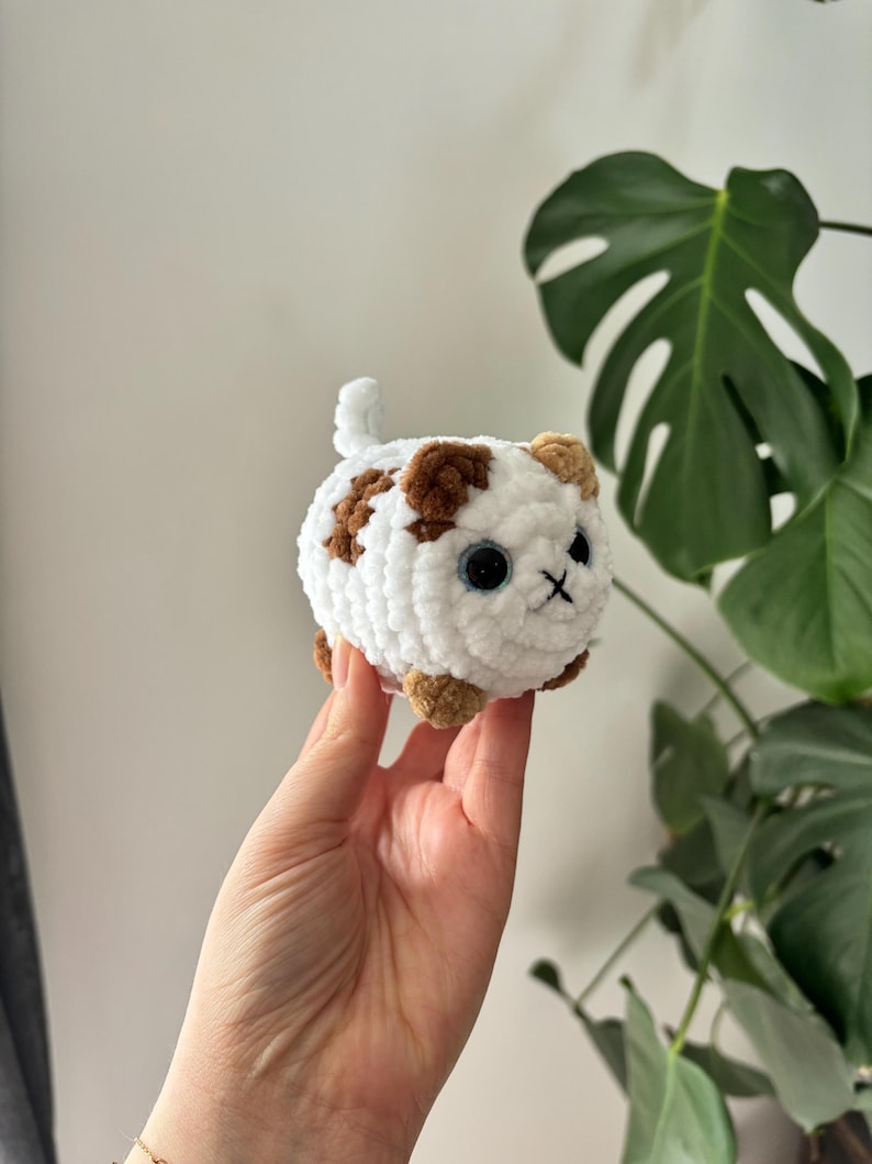 May include: A handmade, white crocheted cat with brown spots, blue eyes, and a stitched black mouth. The small, round cat has tan paws and ears, and a small tail. A cute, soft, and cuddly cat toy.