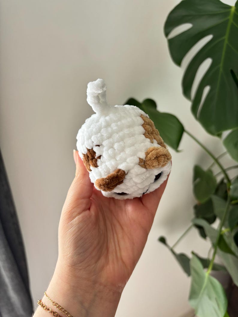 May include: A handmade, plush, white and brown crocheted cat toy. The toy has a rounded body with brown spots and a white tail. The cat toy is held in a hand, with a green plant in the background.