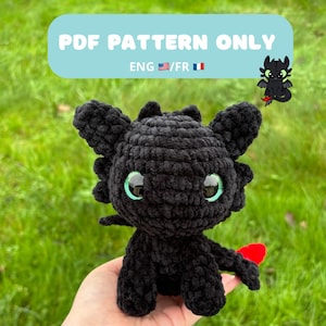 May include: A black crocheted dragon plushie with large green eyes and a red tail fin. The image includes text that reads "PDF PATTERN ONLY" and "ENG /FR". A small cartoon dragon is in the upper right corner.