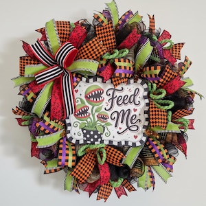 May include: A decorative wreath featuring a central sign with the text Feed Me and illustrations of three green carnivorous plants in a polka dot pot. The wreath is composed of layered ribbons in black, orange, lime green, and purple patterns.