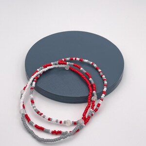 Könnte beinhalten: Drei Perlenarmbänder in den Farben Rot, Weiß und Grau. Die Armbänder sind übereinander gelegt und liegen auf einer blauen Oberfläche.