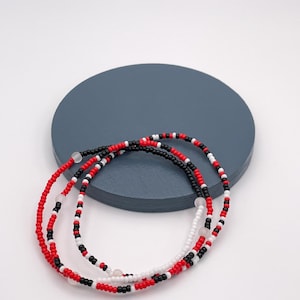 May include: Three beaded bracelets in red, black, and white. The bracelets are layered on top of each other and are on a gray surface.