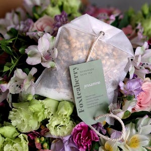May include: A floral arrangement featuring a white bath tea bag with a tag reading "Foxteaberry jessamine." The tag lists ingredients: epsom salt, lemon essential oil, jasmine buds, lemongrass, and raspberry leaves. Flowers in shades of pink, purple, and green.