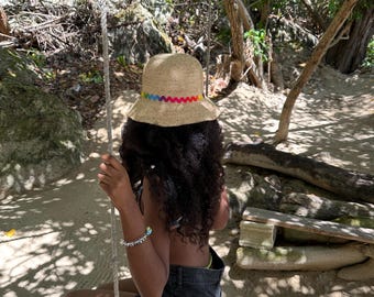 Puede incluir: Una persona con un sombrero de paja con un borde de arcoíris se sienta en un columpio de madera en un entorno de bosque tropical.