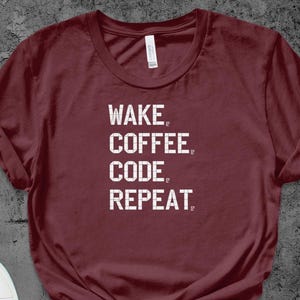Może przedstawiać: Bordowa koszulka z białym napisem "WAKE. COFFEE. CODE. REPEAT." ułożonym pionowo. Koszulka ma okrągły dekolt i krótkie rękawy, z podwiniętymi rękawami.