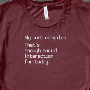 Op de afbeelding: Bordeauxrood T-shirt met de witte tekst: "My code compiles. That's enough social interaction for today." Het shirt heeft een ronde hals en korte mouwen. Een label is zichtbaar bij de halslijn.