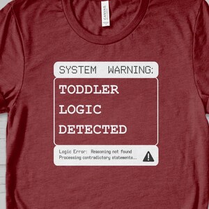 Puede incluir: Una camiseta color granate con un gráfico blanco que dice "SYSTEM WARNING: TODDLER LOGIC DETECTED." Debajo del gráfico hay un mensaje que dice "Logic Error: Reasoning not found. Processing contradictory statements..." con un signo de exclamación.