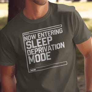 May include: A dark green t-shirt with a white graphic of a computer screen displaying the text "NOW ENTERING SLEEP DEPRIVATION MODE".
