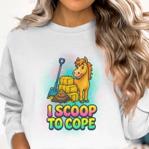 Op de afbeelding: Witte sweatshirt met een cartoon paard, een schop, hooibalen en mest. De tekst "I SCOOP TO COPE" staat in kleurrijke letters. Het ontwerp staat tegen een zachte, pastelkleurige achtergrond.