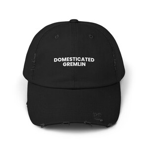 May include: Black distressed baseball cap with the text "DOMESTICATED GREMLIN" in white block letters. The cap has a curved brim and a distressed, worn look.