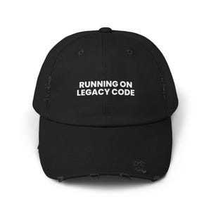 Op de afbeelding: Zwarte baseballcap met een distressed look en de tekst "RUNNING ON LEGACY CODE" in witte blokletters. De cap heeft een gebogen klep en een versleten uitstraling.