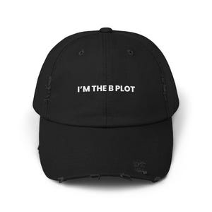 Pode incluir: Boné de beisebol preto desgastado com o texto "I'M THE B PLOT" em branco. O boné tem uma aba curva e um visual desgastado.