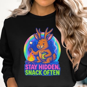 Op de afbeelding: Zwart sweatshirt met een cartoon konijn met geweien dat een marshmallow roostert boven een kampvuur. Het konijn houdt een snackzak vast. De tekst "STAY HIDDEN, SNACK OFTEN" staat eronder. De achtergrond is een regenboog.