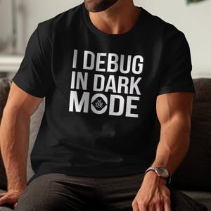 Puede incluir: Una camiseta negra con texto blanco que dice "I DEBUG IN DARK MODE". El texto está centrado en la camiseta y está rodeado de un pequeño icono de error.