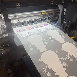 K&ouml;nnte beinhalten: Ein Gro&szlig;formatdrucker druckt eine wei&szlig;e Vinylfolie mit einem sich wiederholenden Muster aus Schneemann-Silhouetten und Schneeflocken. Die Folie wird mit wei&szlig;er Tinte bedruckt.