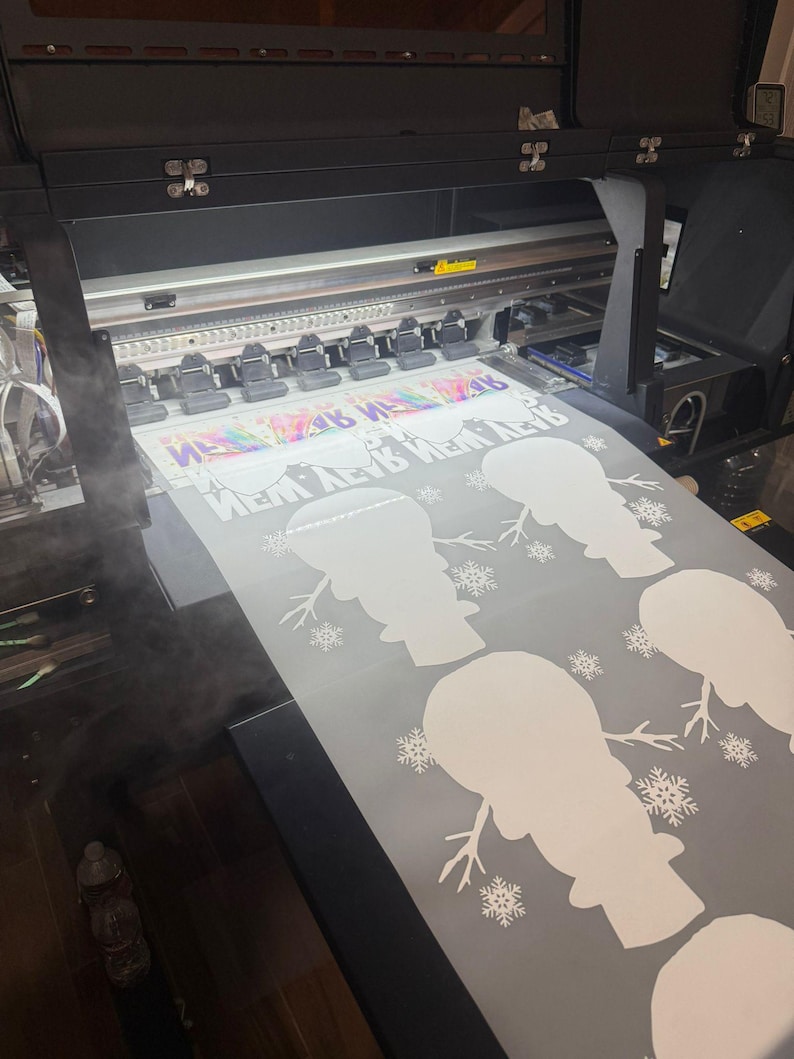 K&ouml;nnte beinhalten: Ein Gro&szlig;formatdrucker druckt eine wei&szlig;e Vinylfolie mit einem sich wiederholenden Muster aus Schneemann-Silhouetten und Schneeflocken. Die Folie wird mit wei&szlig;er Tinte bedruckt.