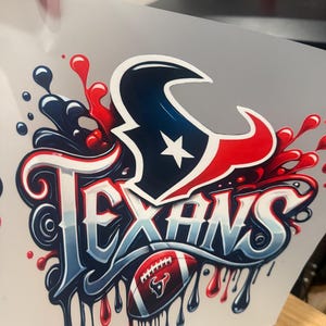K&ouml;nnte beinhalten: Ein wei&szlig;er B&uuml;geltransfer mit einer rot-wei&szlig;-blauen Grafik des Houston Texans Logos mit dem Wort "Texans" in einer stilisierten Schrift. Ein Football mit einem rot-wei&szlig;-blauen Bullenlogo befindet sich unter dem Wort "Texans".