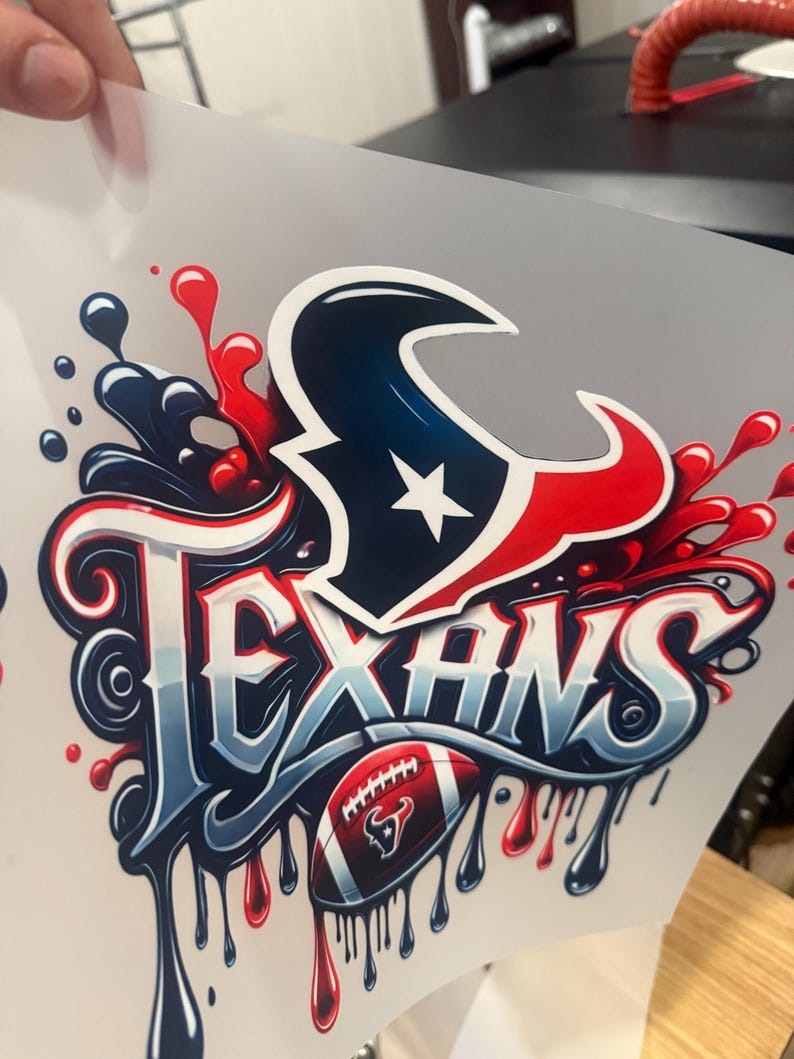 K&ouml;nnte beinhalten: Ein wei&szlig;er B&uuml;geltransfer mit einer rot-wei&szlig;-blauen Grafik des Houston Texans Logos mit dem Wort "Texans" in einer stilisierten Schrift. Ein Football mit einem rot-wei&szlig;-blauen Bullenlogo befindet sich unter dem Wort "Texans".