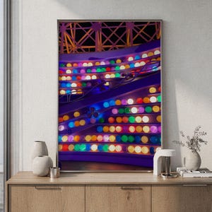Op de afbeelding: Een ingelijste wandkunstprint met een kleurrijke kermisattractie in de nacht. De structuur toont rijen gloeiende, veelkleurige bolvormige lampen in tinten blauw, groen, geel, oranje en roze tegen een donkerpaars frame.