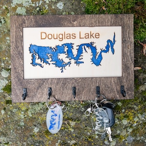 May include: Wooden key holder with a laser-cut map of Douglas Lake. The key holder has four hooks and is stained a dark brown color. The map is blue and white.