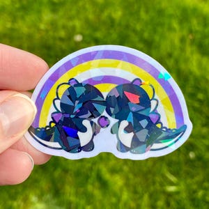 Puede incluir: Una pegatina con dos dinosaurios estilizados abrazándose bajo un arcoíris. Los dinosaurios son de color azul oscuro con patrones geométricos, y el arcoíris tiene secciones amarillas, moradas e iridiscentes. La pegatina se sostiene sobre un fondo verde borroso.