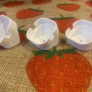 May include: Three white, square-shaped objects with a curved top and a hole in the bottom are displayed. The objects are on a strawberry-patterned fabric. The objects appear to be 3D-printed.