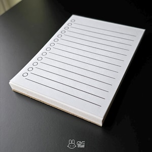 May include: A white notepad with black lines and check circles. The notepad has a light brown edge and is on a black surface. The notepad is for taking notes and making lists.