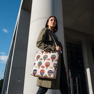 May include: A cream-colored tote bag featuring a repeating pattern of colorful hot air balloons. The balloons are in various colors, including red, blue, and green, with clouds. The bag is carried by a person wearing a green coat and dark jeans.