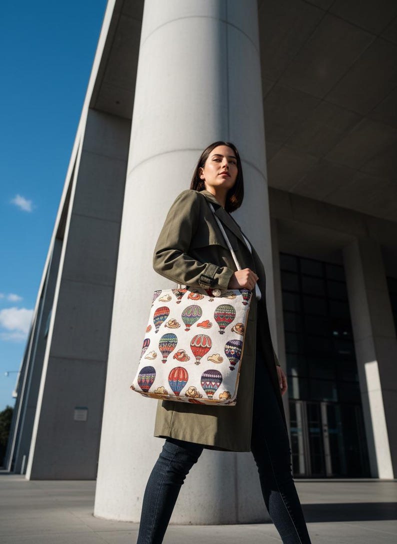 May include: A cream-colored tote bag featuring a repeating pattern of colorful hot air balloons. The balloons are in various colors, including red, blue, and green, with clouds. The bag is carried by a person wearing a green coat and dark jeans.