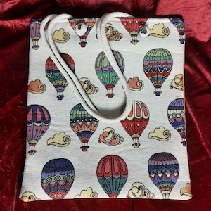 May include: A white tote bag with a repeating pattern of colorful hot air balloons and clouds. The balloons are in various colors, including red, blue, green, and purple. The bag has a white rope handle.