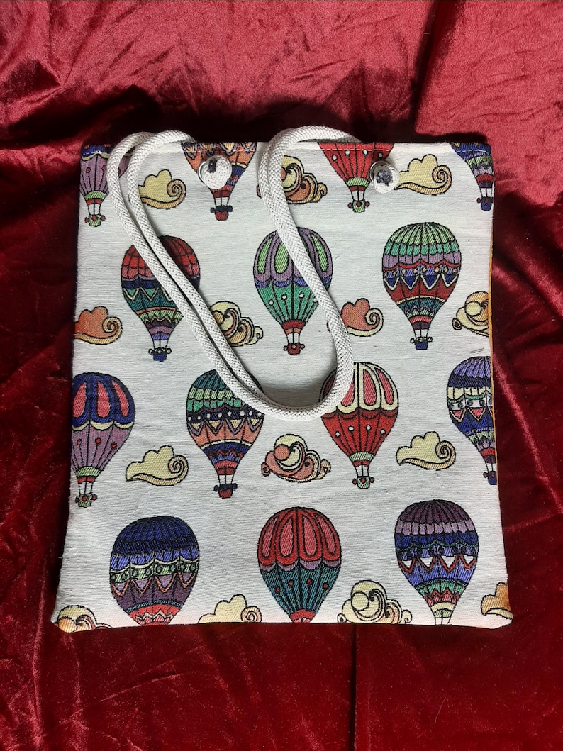 May include: A white tote bag with a repeating pattern of colorful hot air balloons and clouds. The balloons are in various colors, including red, blue, green, and purple. The bag has a white rope handle.