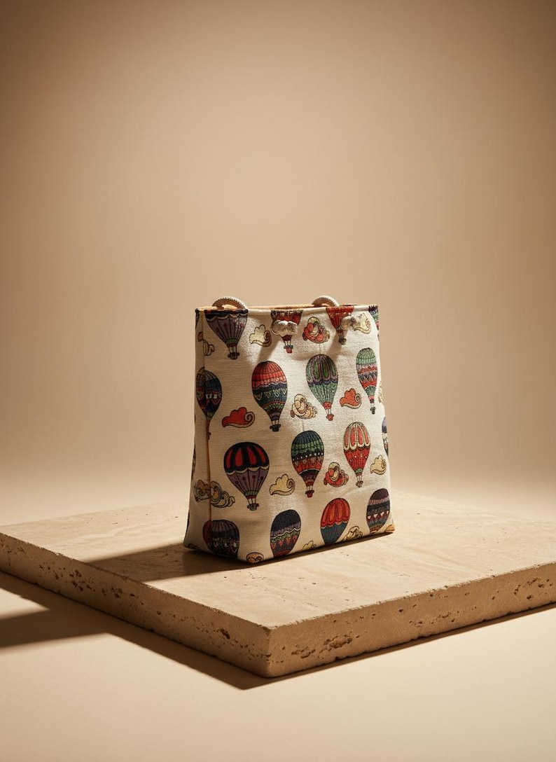 May include: A rectangular tote bag with a cream-colored background and a repeating pattern of colorful hot air balloons. The balloons are red, blue, green, and purple. The bag has rope handles.