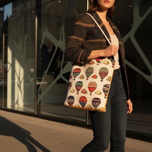 May include: A beige tote bag with a colorful hot air balloon pattern. The bag is carried by a person wearing a striped jacket and dark jeans. The bag has a cream-colored strap and a yellow border.