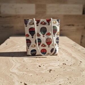 May include: A small tote bag with a repeating pattern of colorful hot air balloons and clouds on a cream background. The bag has a rectangular shape with a white rope handle.